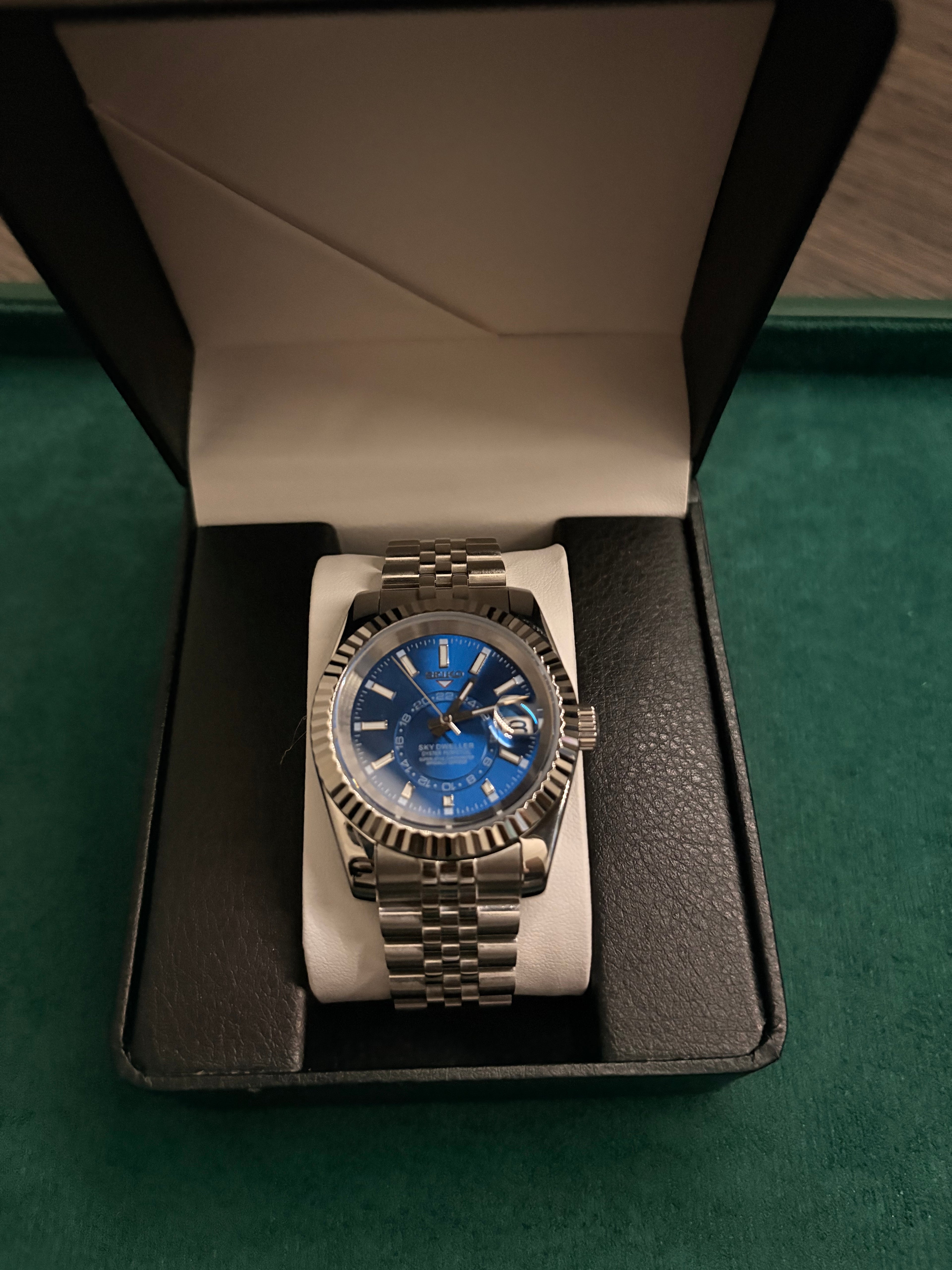 Watch with a blue face and silver case in an open box on a green background