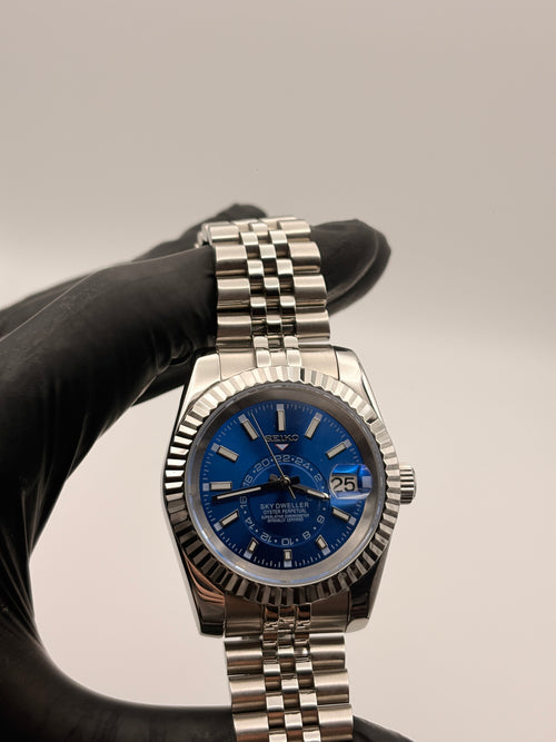 Silver watch with blue face held by a gloved hand against a neutral background