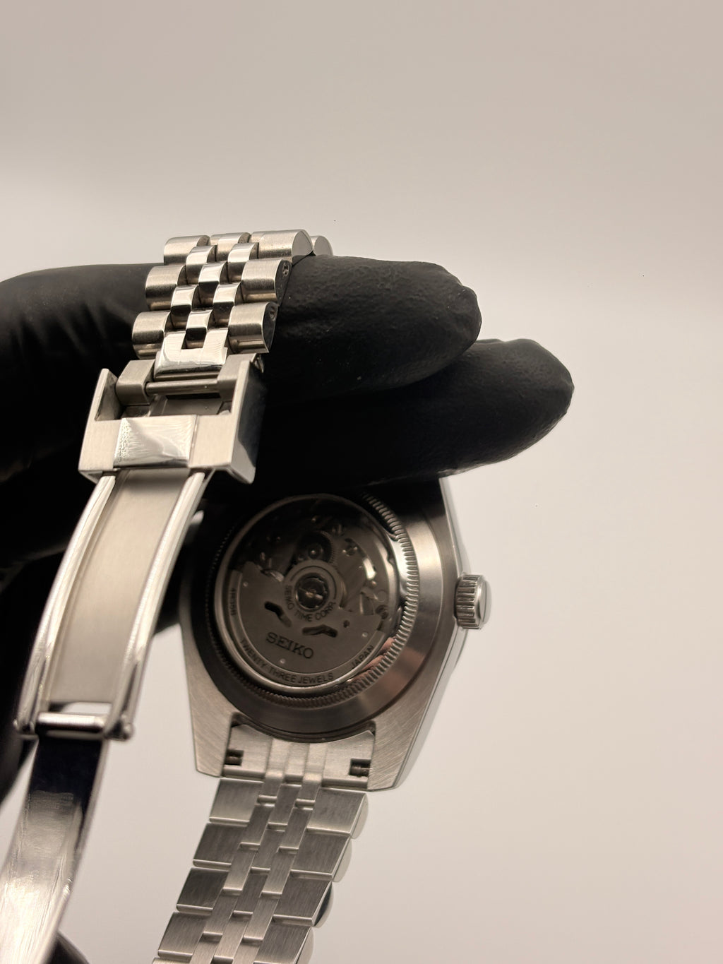 Close-up of a silver watch with a checkered band held by a black-gloved hand against a light gray background.