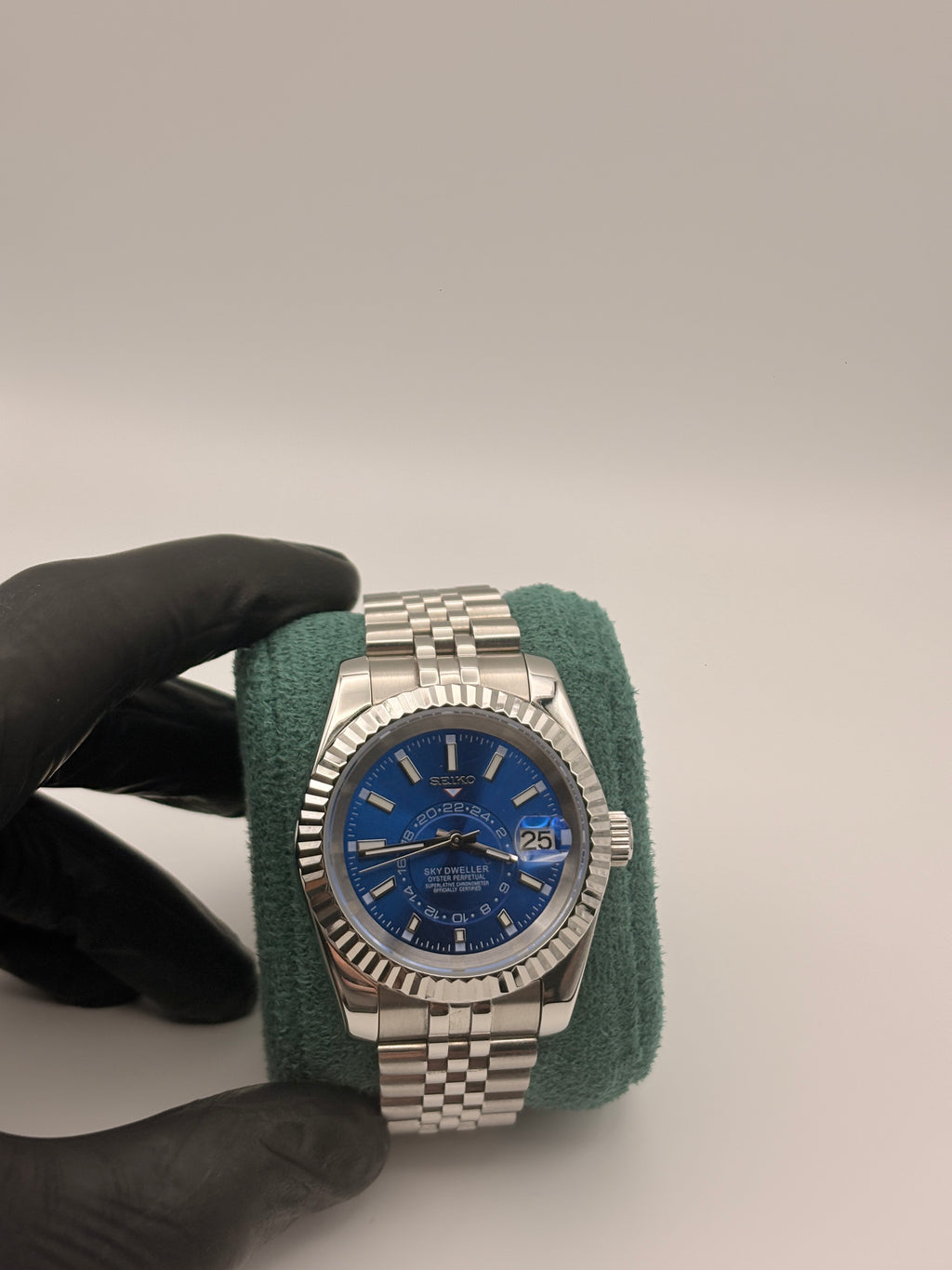 Silver watch with blue face held by a gloved hand against a neutral background
